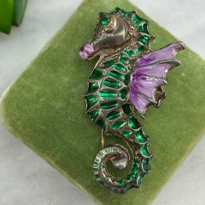 May include: A silver seahorse brooch with green and purple enamel. The seahorse has a detailed design and a curled tail.