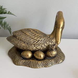 May include: A polished brass sculpture of a mother duck sitting in a nest with two eggs and a small duckling. The metal figure features detailed feather textures and a smooth, reflective surface, depicting the bird in a resting position.