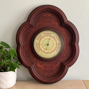 May include: A vintage-style barometer with a wooden frame in a dark red color. The barometer face is white with black markings and has the words "Stormy", "Rain", "Change", "Fair", and "Very Dry" printed around the edge. The frame is intricately carved with a floral design.