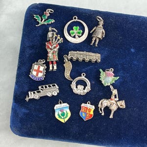 Op de afbeelding: Een verzameling van 13 zilveren bedels met verschillende symbolen van het Verenigd Koninkrijk, waaronder een distel, een klavertjevier, een kroon, een bus en een ridder te paard. De bedels worden getoond op een blauwe fluwelen achtergrond.
