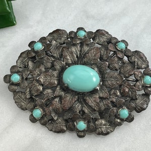 Pode incluir: Um broche de tom prateado com uma grande pedra turquesa em cabochão cercada por pedras turquesas menores engastadas em desenhos florais.