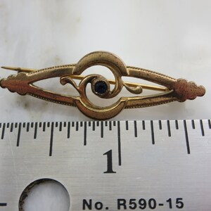 Pode incluir: Um broche dourado com um design intrincado que apresenta uma pedra preciosa azul no centro. O broche mede aproximadamente 2,5 cm de comprimento e tem um pino na parte de tr&aacute;s.
