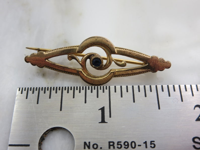 Pode incluir: Um broche dourado com um design intrincado que apresenta uma pedra preciosa azul no centro. O broche mede aproximadamente 2,5 cm de comprimento e tem um pino na parte de tr&aacute;s.