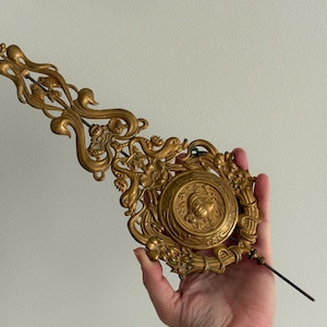 May include: Ornate gold-colored clock pendulum with an elaborate design. The pendulum features a central medallion with a detailed relief and is held by a hand. The decorative piece has a long, slender rod and is likely a vintage or antique item.