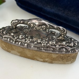 Op de afbeelding: Een antieke verzilverde kledingborstel met een sierlijk handvat en bloemendetails. De borstel heeft een rechthoekige vorm met een lichtbruine borstelbasis. De handgreep is gebogen en heeft ingewikkelde krullen. De borstel is waarschijnlijk uit de vroege 20e eeuw.