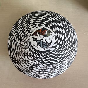Puede incluir: Un cuenco redondo de cerámica en blanco y negro con un diseño geométrico en espiral. El centro presenta un diseño circular con dos figuras estilizadas de peces y lagartos en tonos marrones, verdes y grises. El cuenco es una pieza decorativa.