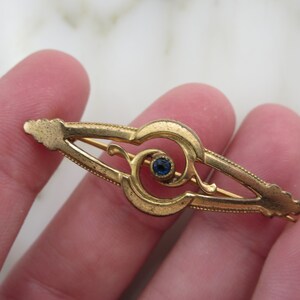 Pode incluir: Um broche dourado com uma pedra preciosa azul no centro. O broche tem a forma de um redemoinho estilizado.