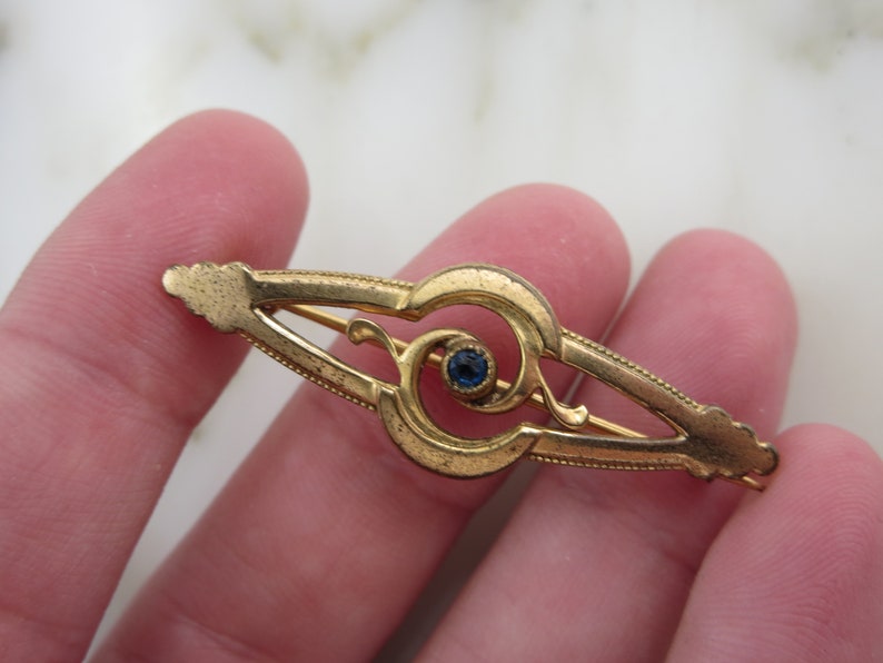 Pode incluir: Um broche dourado com uma pedra preciosa azul no centro. O broche tem a forma de um redemoinho estilizado.