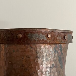 May include: Close-up of the top of a hammered copper pitcher with a rusty iron band and rivets around the rim.
