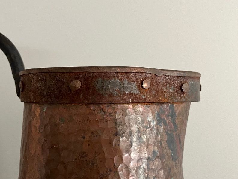 May include: Close-up of the top of a hammered copper pitcher with a rusty iron band and rivets around the rim.