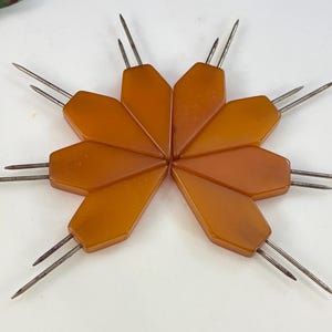 May include: A collection of vintage amber-colored hat pins arranged in a starburst pattern. Each pin features a geometric, faceted head and two silver-toned needles. The pins are displayed on a white surface.