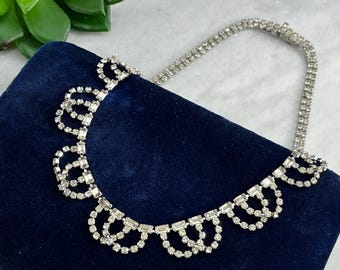 May include: A silver rhinestone necklace with a delicate, scalloped design. The necklace is made of small, clear rhinestones that are set in a silver metal setting. The necklace is on a dark blue velvet background.