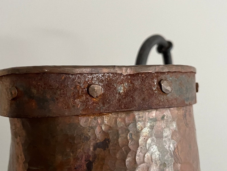 May include: A rustic copper bucket with a hammered finish and a black metal handle. The bucket has a metal band around the top with decorative rivets.