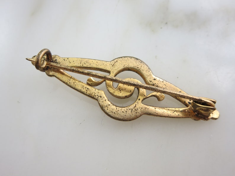 Pode incluir: Broche de clave de sol dourada com um design decorativo. A broche tem um fecho de pino e fecho.
