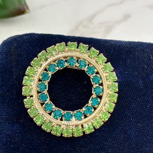 May include: A circular brooch featuring two concentric rings of faceted rhinestones. The outer ring consists of lime green stones, while the inner ring features teal stones. A gold-toned twisted rope metal border separates the two color rows.