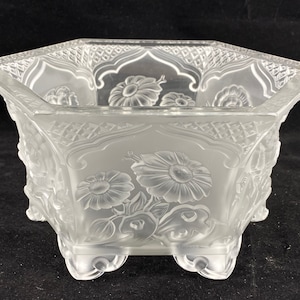 Puede incluir: Un cuenco de vidrio transparente con forma hexagonal y acabado esmerilado. El cuenco tiene un intrincado diseño floral grabado en la superficie y descansa sobre cuatro patas.