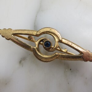 Pode incluir: Um broche dourado com uma pedra preciosa azul no centro. O broche tem a forma de um olho estilizado com detalhes intrincados.