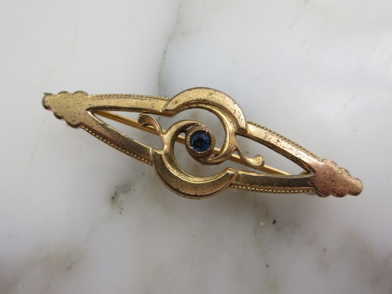 Pode incluir: Um broche dourado com uma pedra preciosa azul no centro. O broche tem a forma de um olho estilizado com detalhes intrincados.