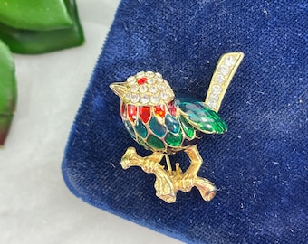 May include: A gold-tone brooch featuring a green, red, and blue bird perched on a branch. The bird is decorated with clear and red rhinestones.