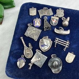 May include: A collection of vintage silver charm pendants on a blue velvet background. The charms feature various designs, including a canoe, a bench, a graduation cap, a cowboy hat, a tennis racket, and several city and state emblems.