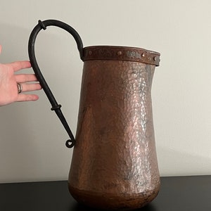 May include: A hammered copper pitcher with a black wrought iron handle. The pitcher has a wide mouth and a rounded body.