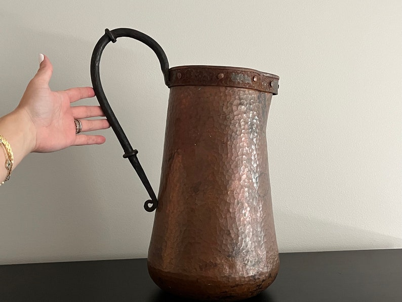 May include: A hammered copper pitcher with a black wrought iron handle. The pitcher has a wide mouth and a rounded body.
