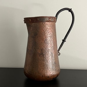 May include: A hammered copper pitcher with a black iron handle. The pitcher has a wide mouth and a rounded body.
