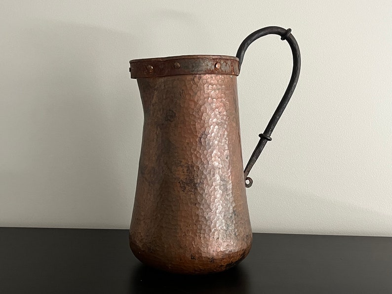 May include: A hammered copper pitcher with a black iron handle. The pitcher has a wide mouth and a rounded body.