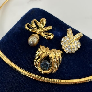May include: A gold-tone omega chain necklace paired with three distinct pendants: one featuring a gold bow with a dangling pearl, one with a central dark stone surrounded by gold swirls, and one encrusted with clear crystals in a cross pattern.
