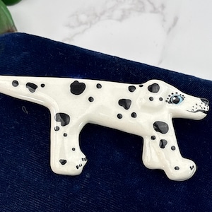 May include: A ceramic Dalmatian dog figurine. The dog is white with black spots and has blue eyes. The figurine is lying on a dark blue surface. The dog's head is turned to the right.