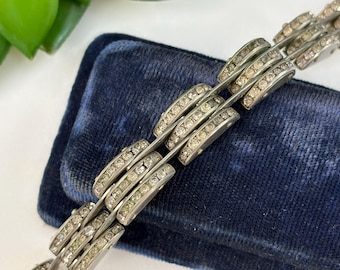 May include: Silver-toned bracelet with a repeating design of curved links, each encrusted with small, clear rhinestones. The bracelet is displayed on a dark blue velvet surface, with a hint of green foliage in the background. A piece of jewelry.