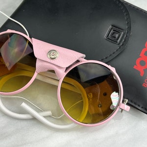 Könnte beinhalten: Rosa runde Sonnenbrille mit braunen Gläsern und einem weißen Band. Die Sonnenbrille ist zusammengeklappt und liegt auf einem schwarzen Lederetui mit dem in Rot gedruckten Text "Jones Boulder Colorado".