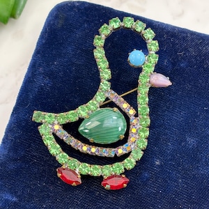 May include: A vintage duck brooch with green, red, and blue rhinestones. The duck has a large green stone in the center, a blue stone for the eye, and a pink stone for the beak. The brooch is set on a dark blue velvet surface.