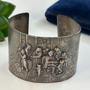 May include: Silver-toned cuff bracelet with a detailed relief scene of figures in a room. The bracelet has a wide band with a rectangular shape and a slightly curved design. The relief depicts people, a dog, and a barrel.