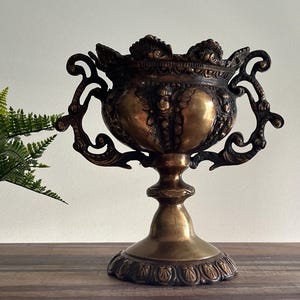 May include: Ornate gold-toned metal vase with two handles and intricate detailing. The vase has a round body and a pedestal base.