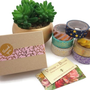 Pode incluir: Uma pequena caixa de cart&atilde;o com um design estampado em rosa e dourado e um adesivo dourado que diz "obrigado". A caixa est&aacute; sobre uma superf&iacute;cie branca ao lado de uma pilha de fitas washi coloridas e um cart&atilde;o de visita que diz "Vintage in Bloom".