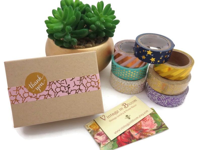 Pode incluir: Uma pequena caixa de cart&atilde;o com um design estampado em rosa e dourado e um adesivo dourado que diz "obrigado". A caixa est&aacute; sobre uma superf&iacute;cie branca ao lado de uma pilha de fitas washi coloridas e um cart&atilde;o de visita que diz "Vintage in Bloom".
