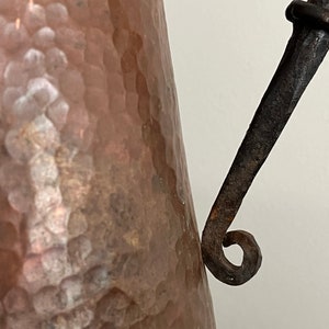 May include: A hammered copper pitcher with a rustic iron handle. The handle is curved and has a decorative loop at the end.