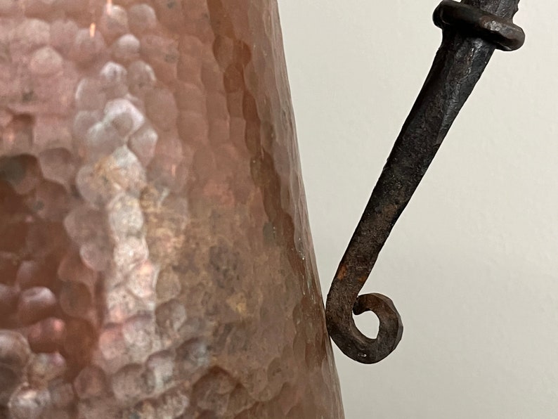 May include: A hammered copper pitcher with a rustic iron handle. The handle is curved and has a decorative loop at the end.