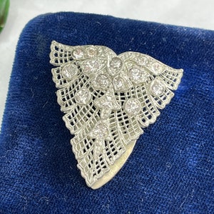 Könnte beinhalten: Eine silberfarbene Brosche aus filigranem Metall mit dreieckiger Form und einem Design aus Blättern und Blüten. Die Brosche ist mit klaren Strasssteinen verziert.