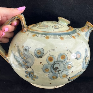 May include: A hand-painted ceramic teapot with a round body and a lid. The teapot is decorated with blue and brown floral designs and birds. The teapot is a traditional Mexican style.