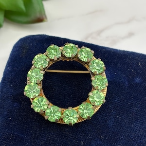 May include: A circular brooch featuring a gold-tone metal frame set with a ring of faceted light green stones. The metal base has a textured, sunburst-like pattern radiating from the center, and a horizontal pin mechanism is visible on the back.
