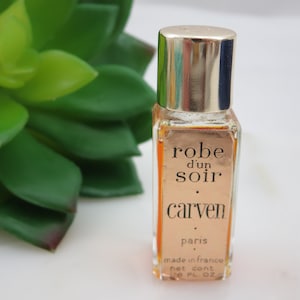 Puede incluir: Un pequeño frasco de vidrio transparente de perfume Carven Robe d'un Soir. El frasco tiene una tapa dorada y una etiqueta con el nombre del perfume en letras negras. El frasco está sobre una superficie blanca.