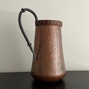 May include: A hammered copper pitcher with a black wrought iron handle. The pitcher has a rustic, aged look and is perfect for adding a touch of vintage charm to your home decor.
