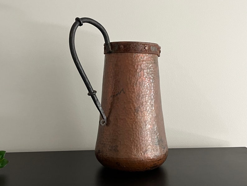 May include: A hammered copper pitcher with a black wrought iron handle. The pitcher has a rustic, aged look and is perfect for adding a touch of vintage charm to your home decor.