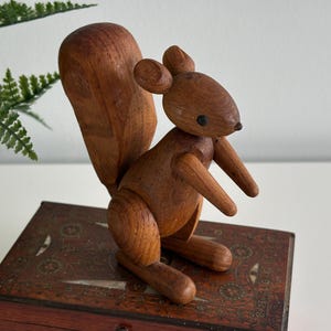 May include: A wooden squirrel ornament with a large tail, small ears, and black eyes. The squirrel is made of light brown wood and is posed in a sitting position. It is displayed on a wooden box with decorative inlaid details.
