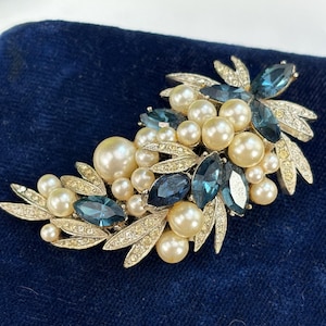 May include: A vintage brooch featuring a floral design with faux pearls and blue-toned gemstones. The brooch has a silver-tone metal setting and is displayed on a dark blue velvet surface. The brooch is a classic piece of jewelry.