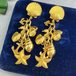 May include: Gold tone earrings with a seashell charm and other charms, including anchors, seahorses, and starfish. The earrings are on a blue velvet background.