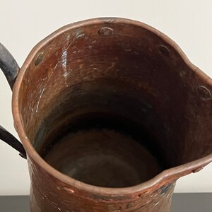 May include: A copper pitcher with a hammered finish and a black iron handle. The pitcher has a wide mouth and a rounded body. The interior of the pitcher is visible and appears to be clean.