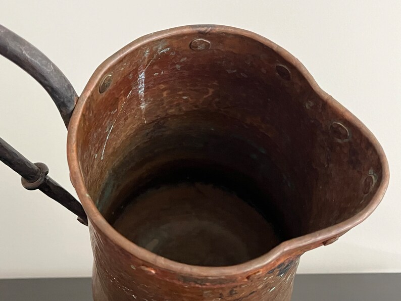 May include: A copper pitcher with a hammered finish and a black iron handle. The pitcher has a wide mouth and a rounded body. The interior of the pitcher is visible and appears to be clean.